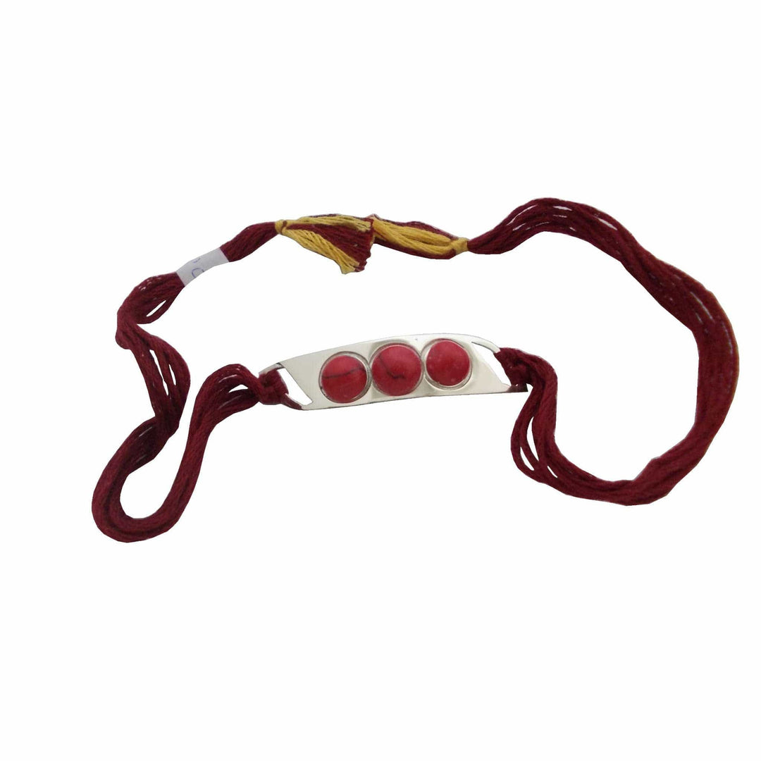 Rakhi Ruby is a silver rakhi with three red stones on a curved bar on a maroon and gold tasselled thread. - white background