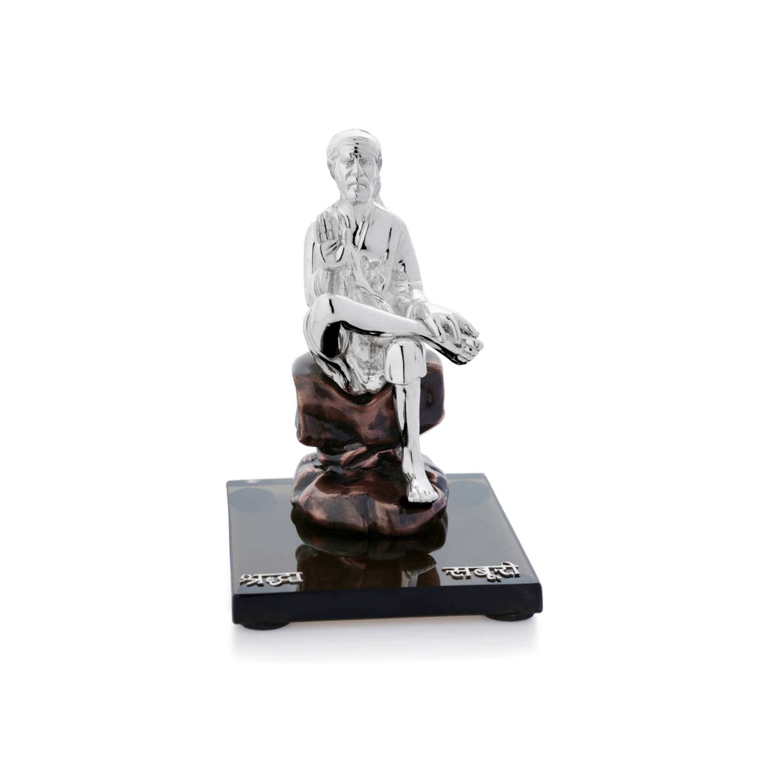 Sai Baba Sitting is a sterling silver idol of Sai Baba sitting cross-legged on a textured bronze-toned rock, one hand raised in blessing - white background
