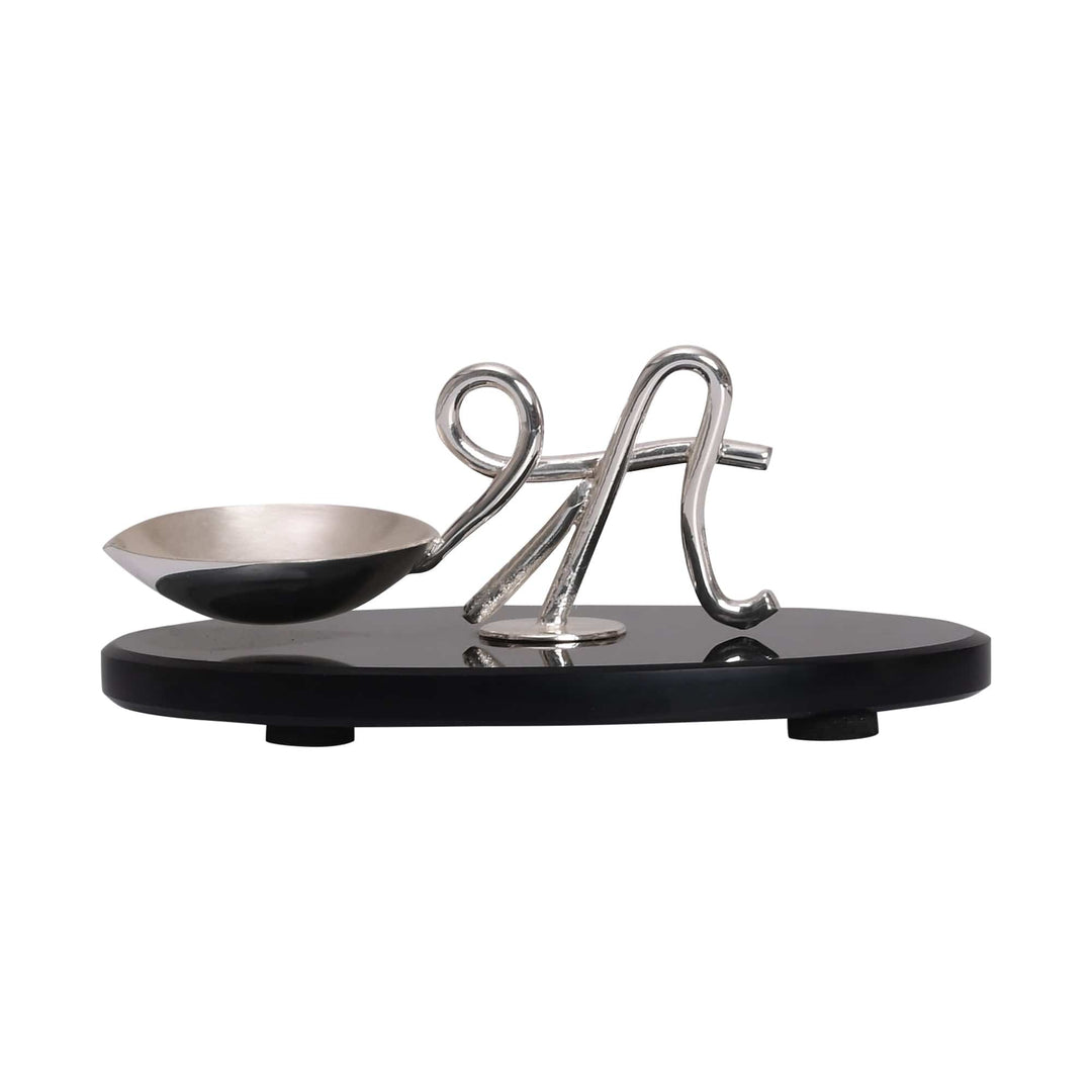 Shree Diya is a silver diya that pairs a small oil cup with a sculptural flourish on a glossy black oval base - white background