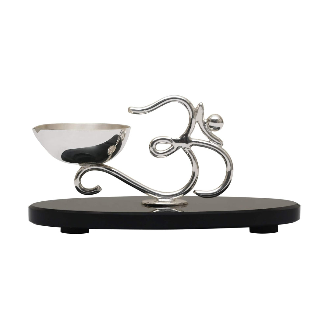 Om Diya On Base pairs a silver diya cup with a sculptural Om symbol rendered in smooth, scrolling silver lines - white background