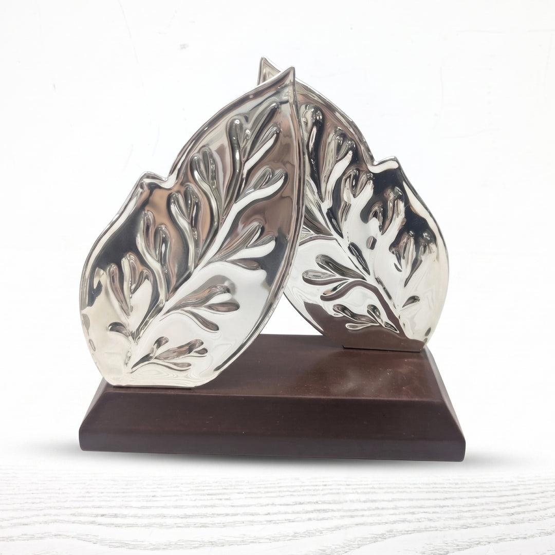 Leaf Tissue Holder is a silver tissue holder with two sculpted leaf panels leaning together on a bevelled dark wood base. - white background